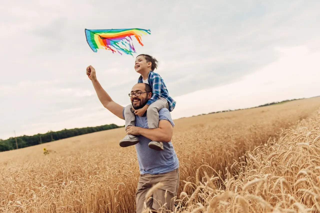 A father carrying his son on his shoulders flying a kite in a wheat field